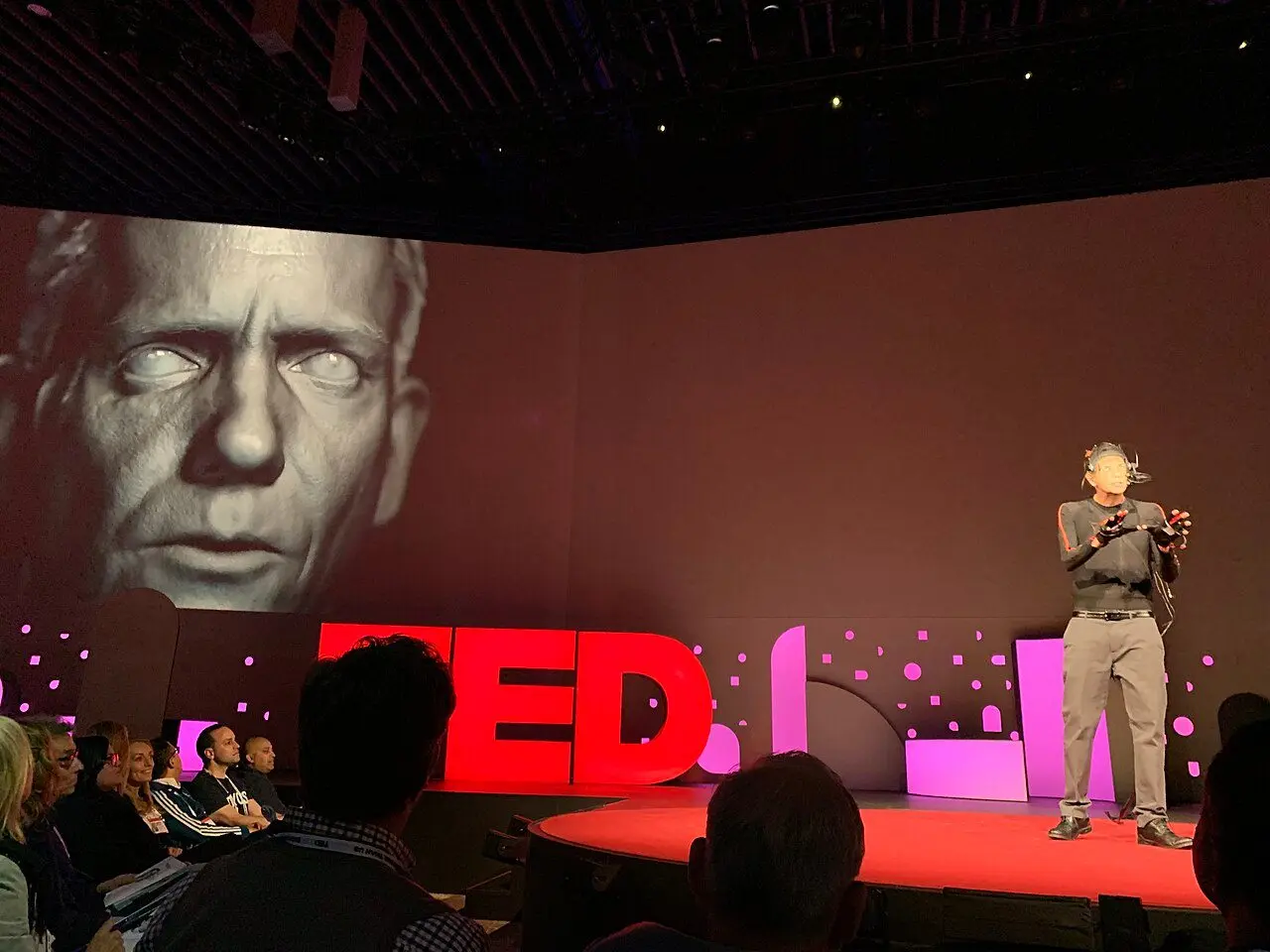 Doug Roble on the TED stage in 2019, wearing motion-capture gear, with the digital face DigiDoug on the screen behind him