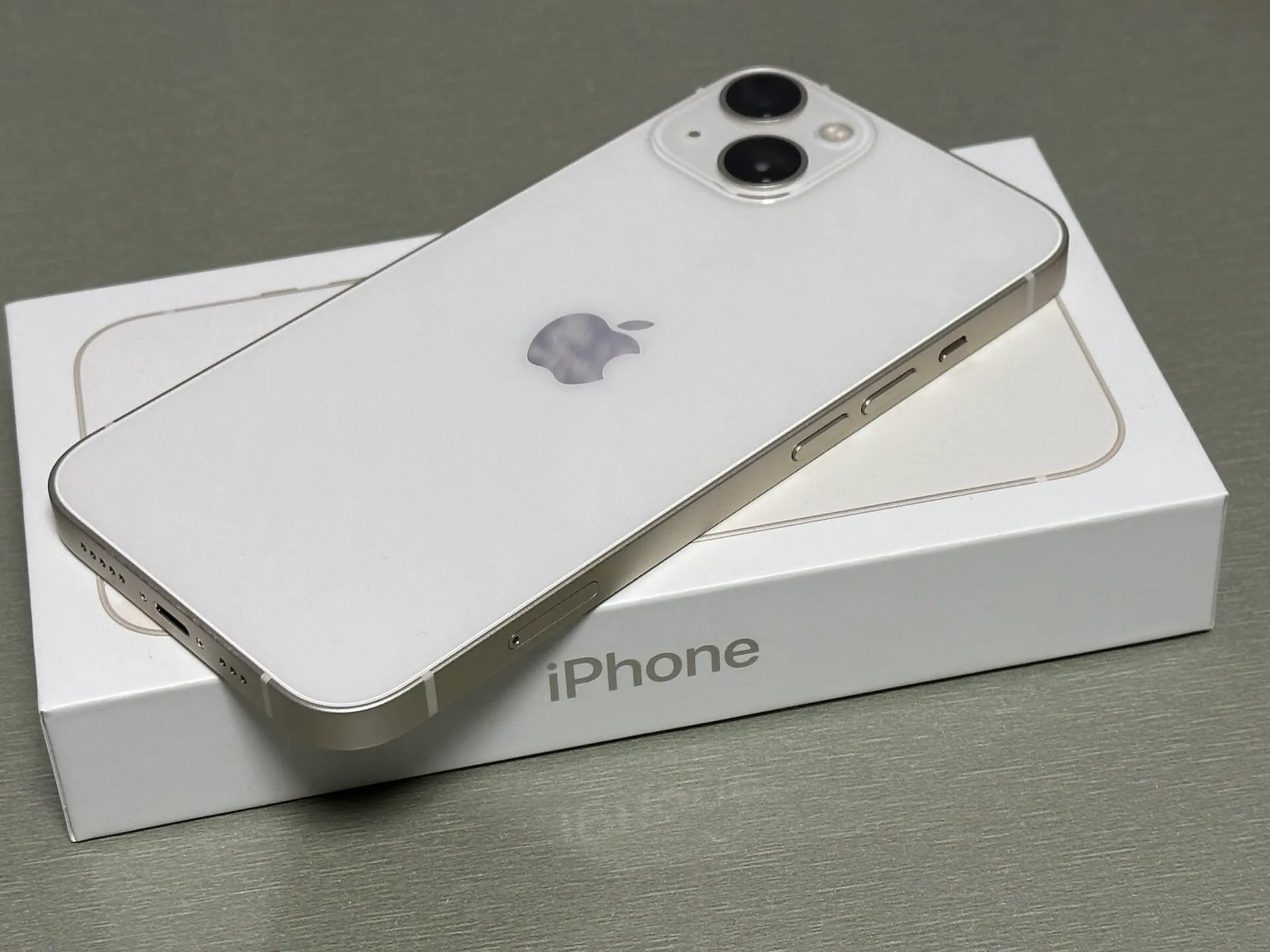 白い化粧箱の上に斜めに置かれたスターライト色の iPhone 13 の背面。2つのカメラが対角に配置されている