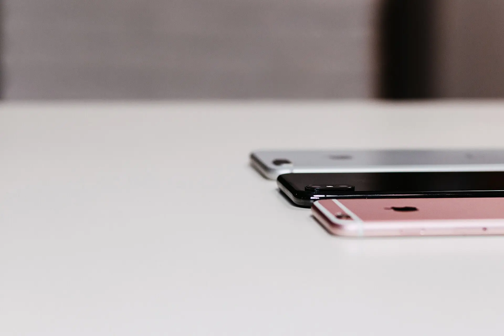 Three iPhones stacked with a slight offset on a white desk, seen from a low angle: the rose-gold iPhone 6s in front, with an iPhone 8 Plus and an iPhone X behind it