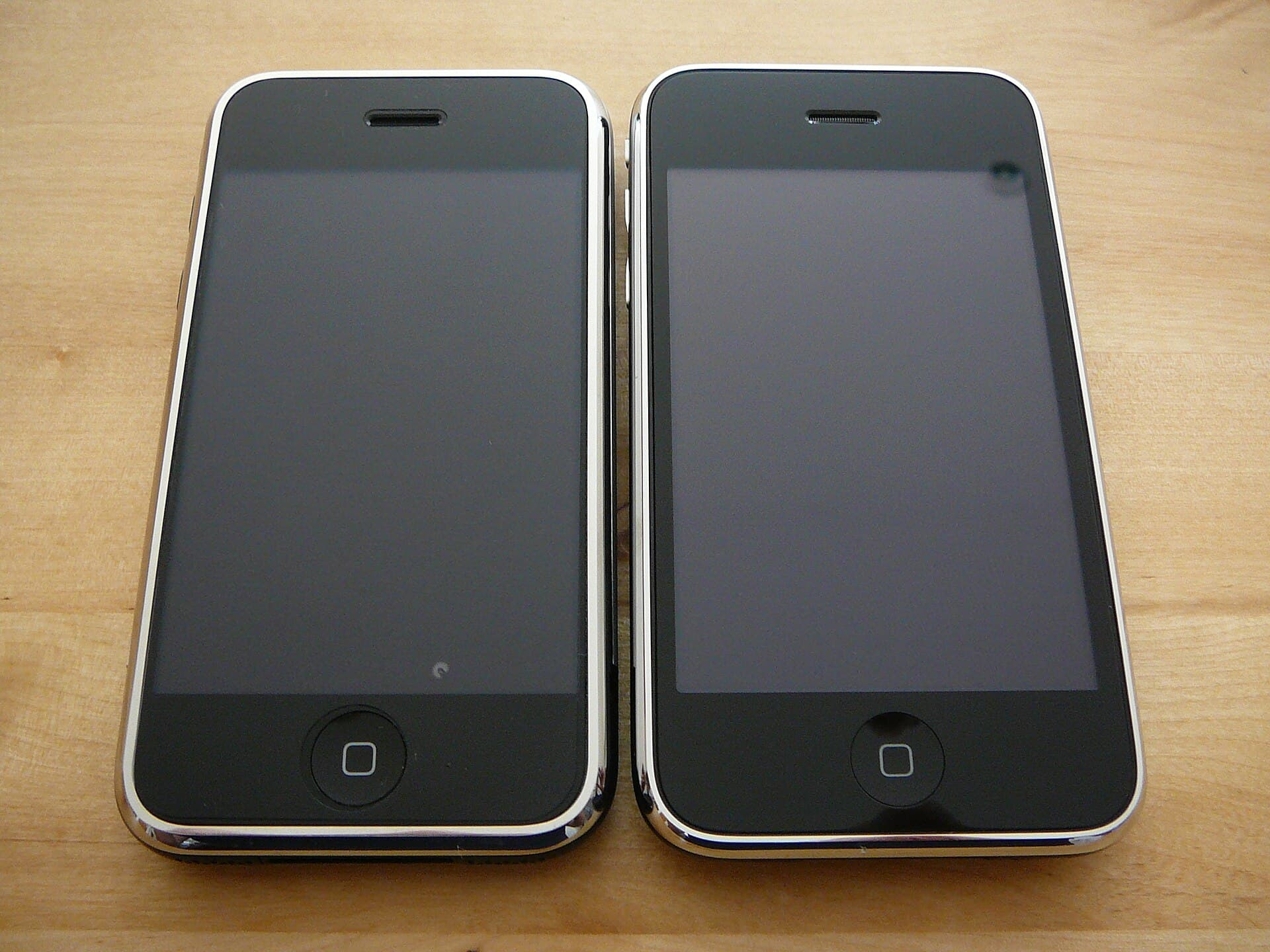 The original iPhone (left) and the iPhone 3G (right) lying front-up side by side on a wooden desk, photographed on the launch day of 11 July 2008
