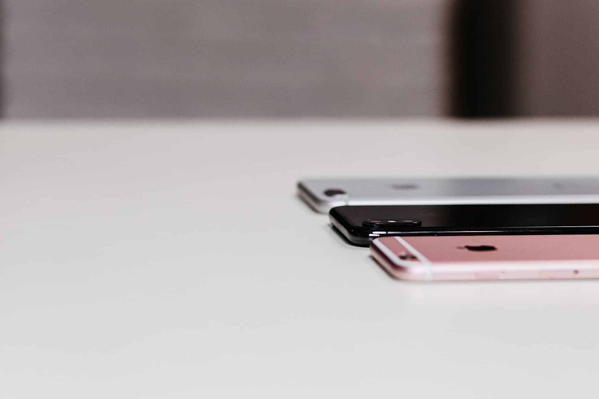 Three iPhones stacked with a slight offset on a white desk, seen from a low angle: the rose-gold iPhone 6s in front, with an iPhone 8 Plus and an iPhone X behind it